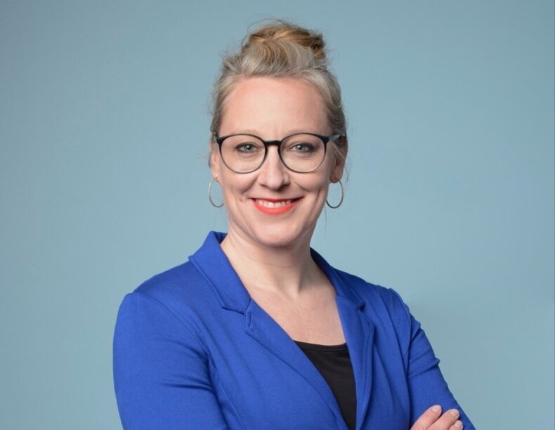 Porträtfoto von Aline Trede in einem blauen Blazer vor hellblauem Hintergrund. Aline Trede verschränkt die Arme.